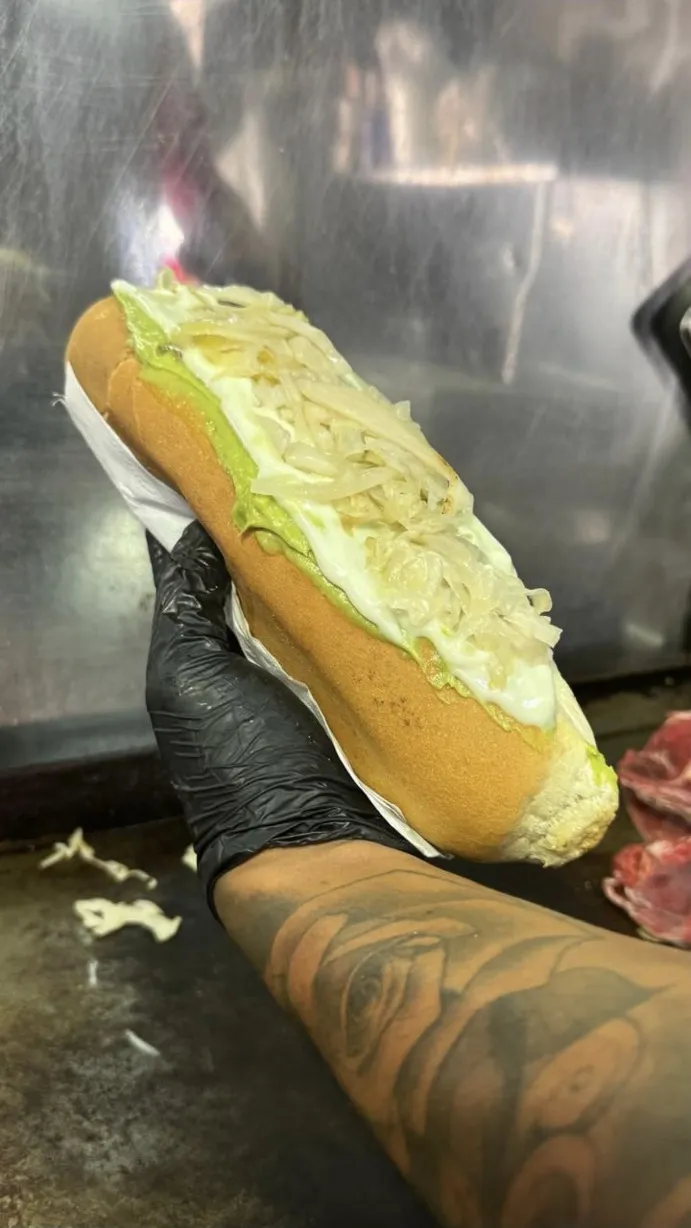 Completo XL recién armado con palta, mayo y chucrut, sostenido con guante sobre la plancha del local.
